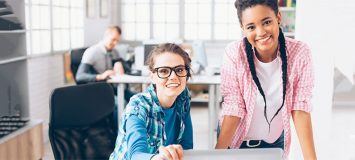 两位软件专业人员在办公桌-右方妇女坐立,左方妇女站立都看着摄像头找新生涯 启动软件质量保证证书课程SAQ101
